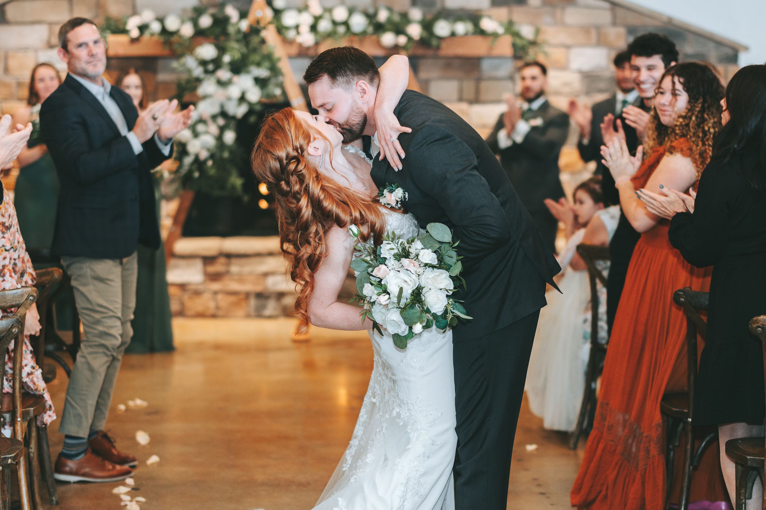 A romantic wedding celebration moment with guests watching as a couple shares a dip kiss beneath floral decor.