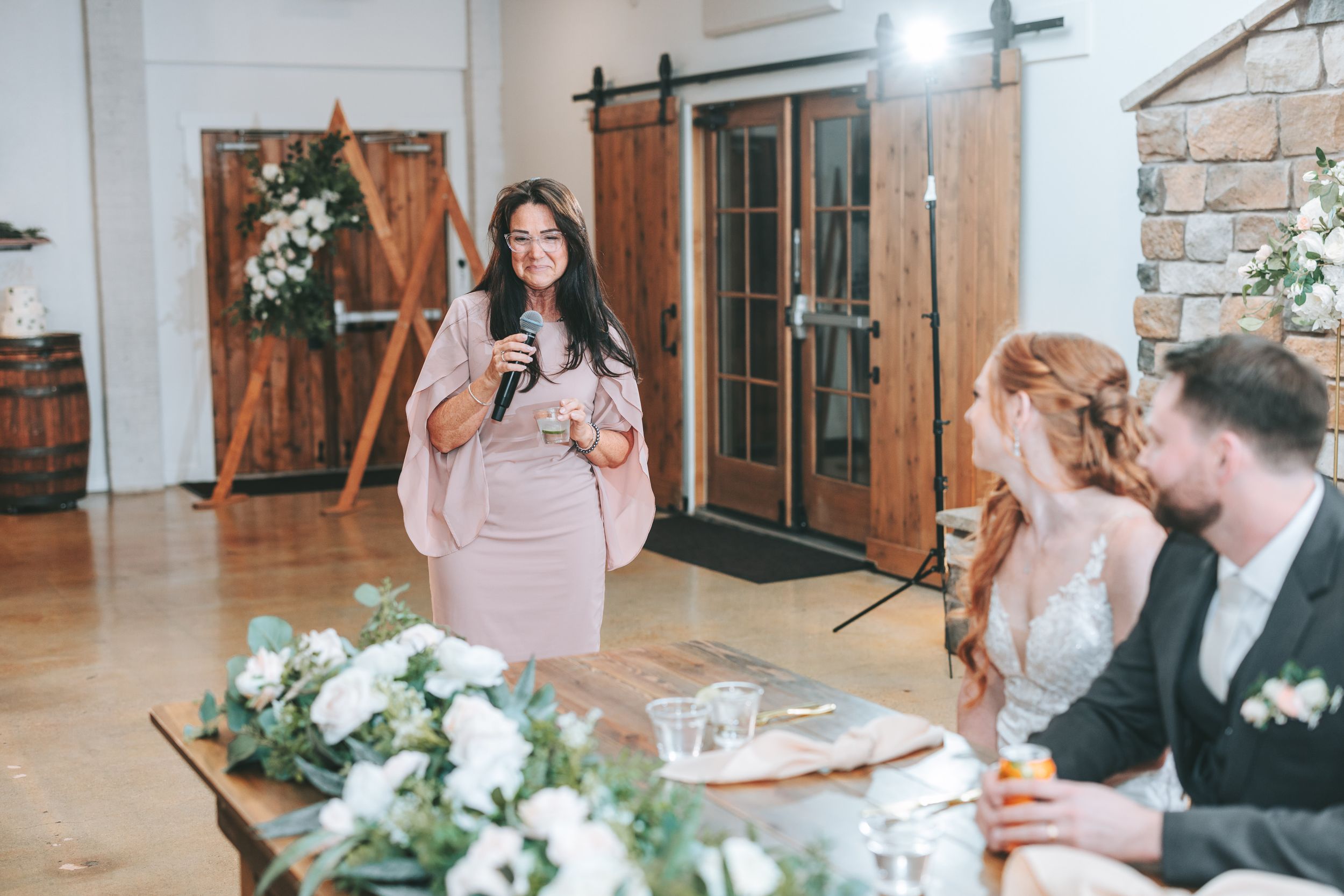 A wedding reception speech is being delivered at a rustic venue with wooden barn doors and elegant white floral arrangements.