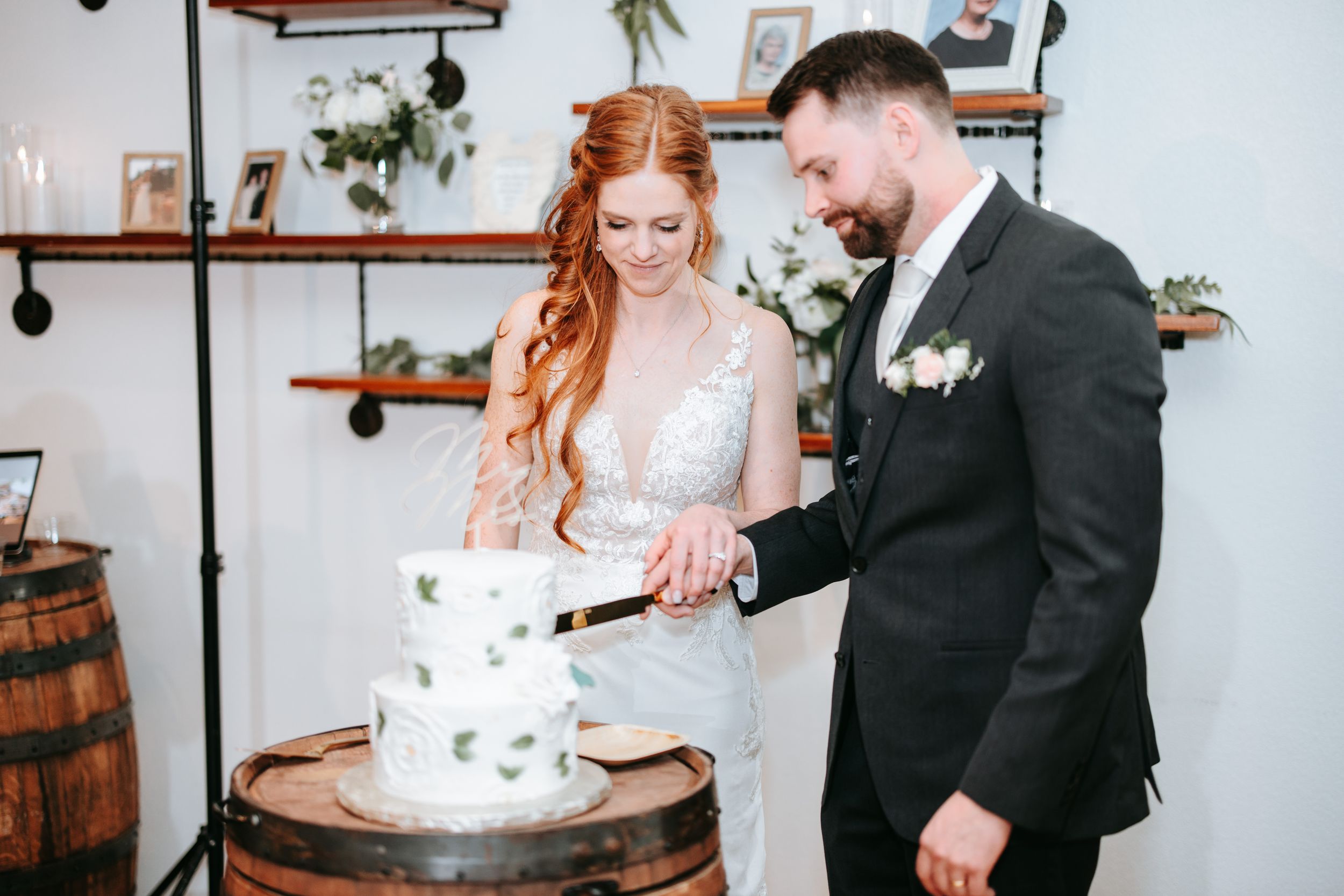 A couple in formal wedding attire smiles while cutting their white wedding cake during their reception celebration.