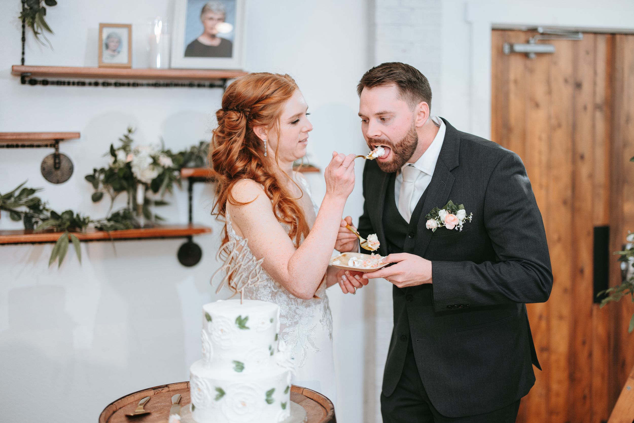 A wedding couple shares a playful moment while feeding each other cake in a rustic indoor venue.