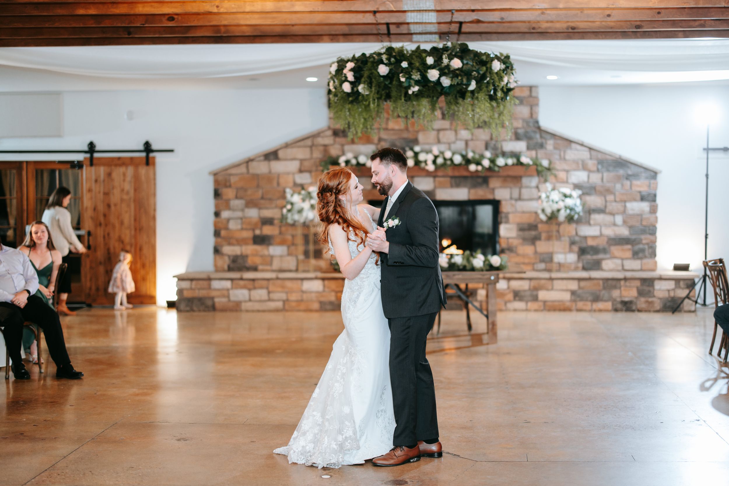 A newlywed couple shares their first dance under floral decorations in an elegant barn-style reception venue.