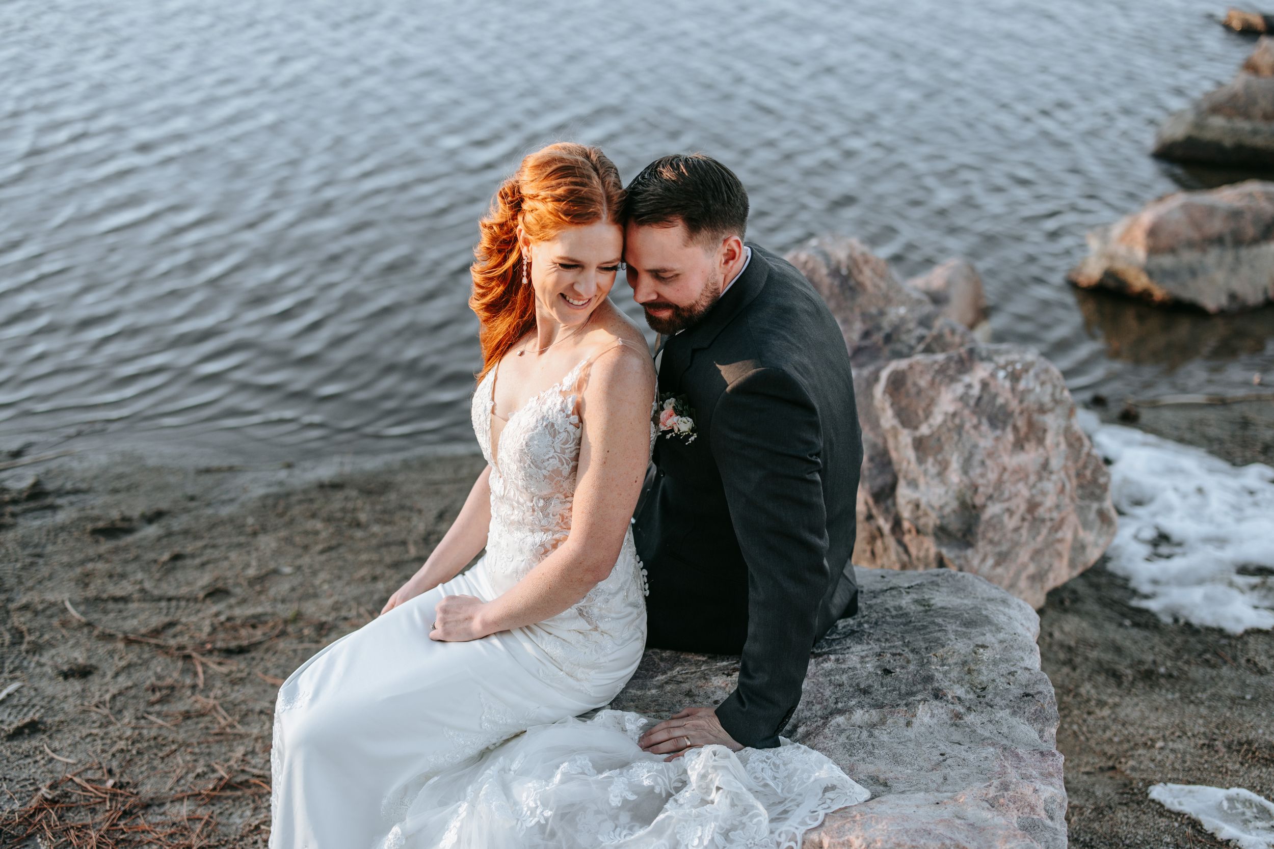 A couple sits intimately together on rocks by a tranquil lake during their wedding photoshoot.
