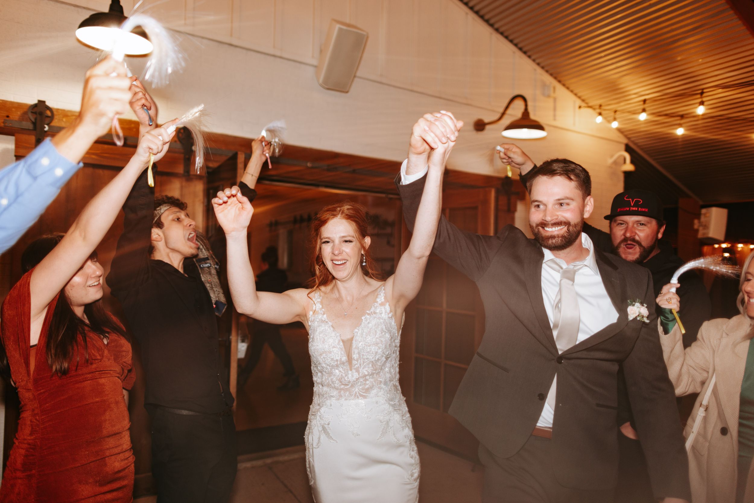 Wedding party dancing and waving napkins joyfully during wedding reception under wooden ceiling with string lights.