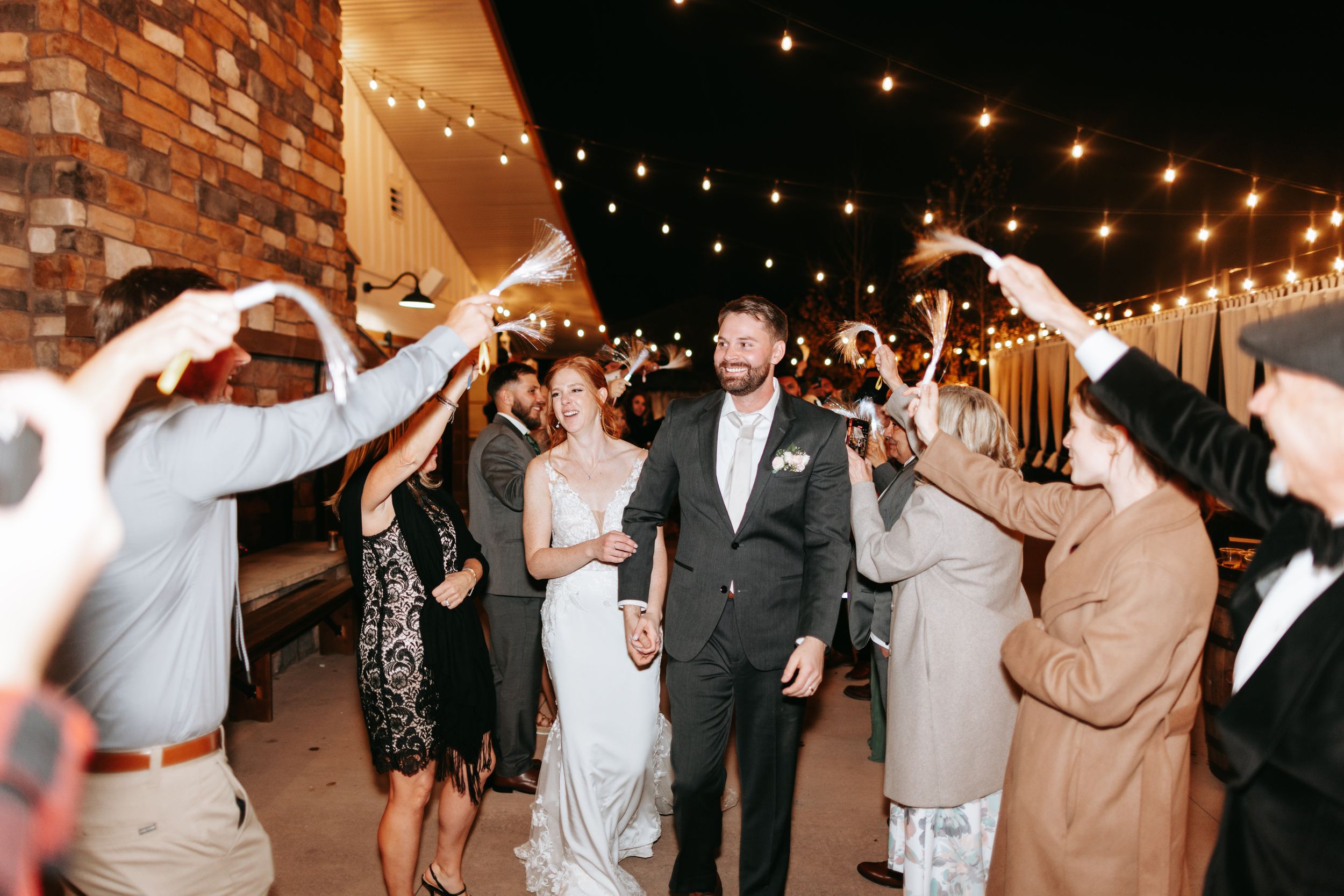 Wedding celebration indoors at night with string lights overhead and guests forming an arch pathway.