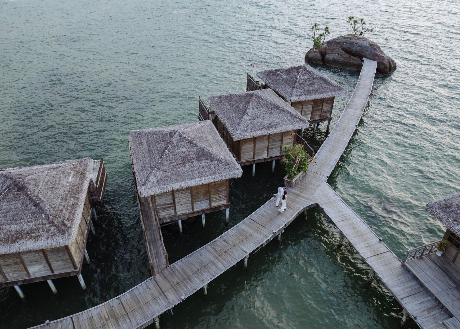 Aerial view of overwater bungalows connected by wooden boardwalk over calm turquoise waters at tropical resort.