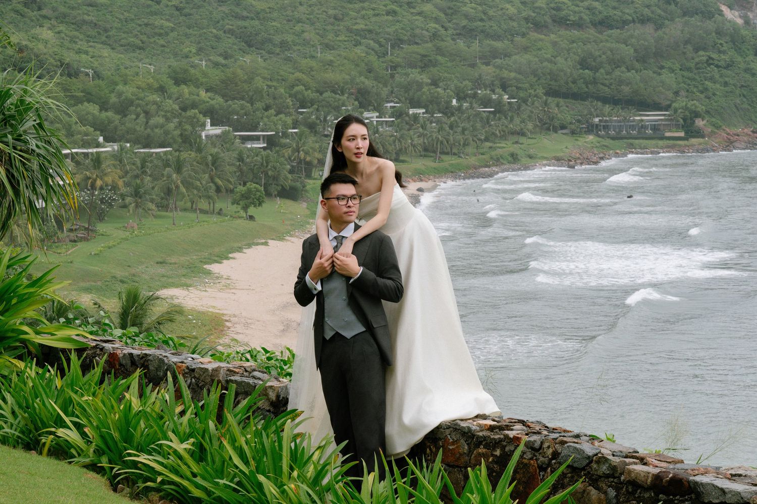 Bride and groom pose dramatically on rocky coastal overlook with stunning panoramic ocean and mountain views behind them.