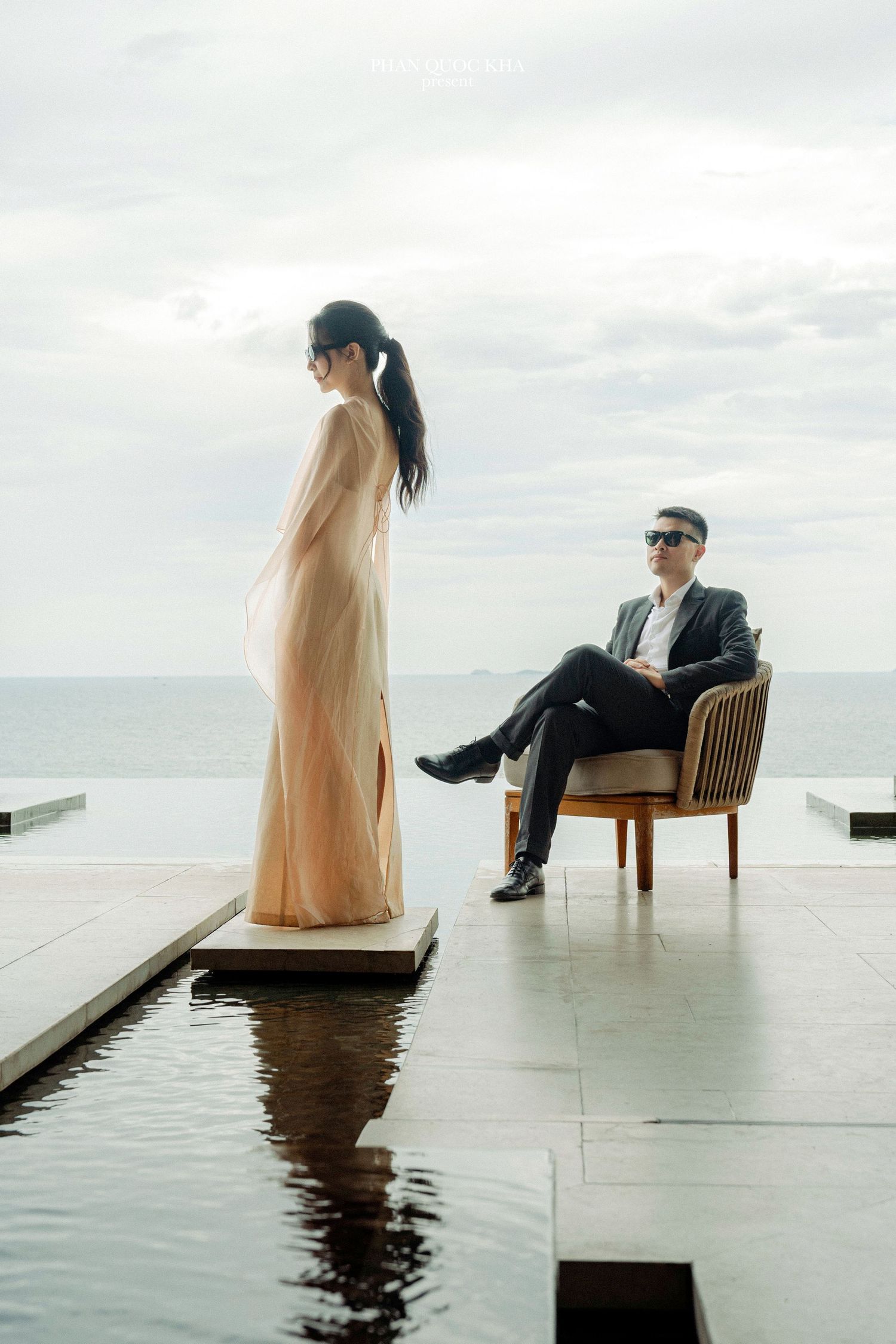 Elegant couple posing by infinity pool overlooking ocean, woman in gold gown, man in suit seated in chair.