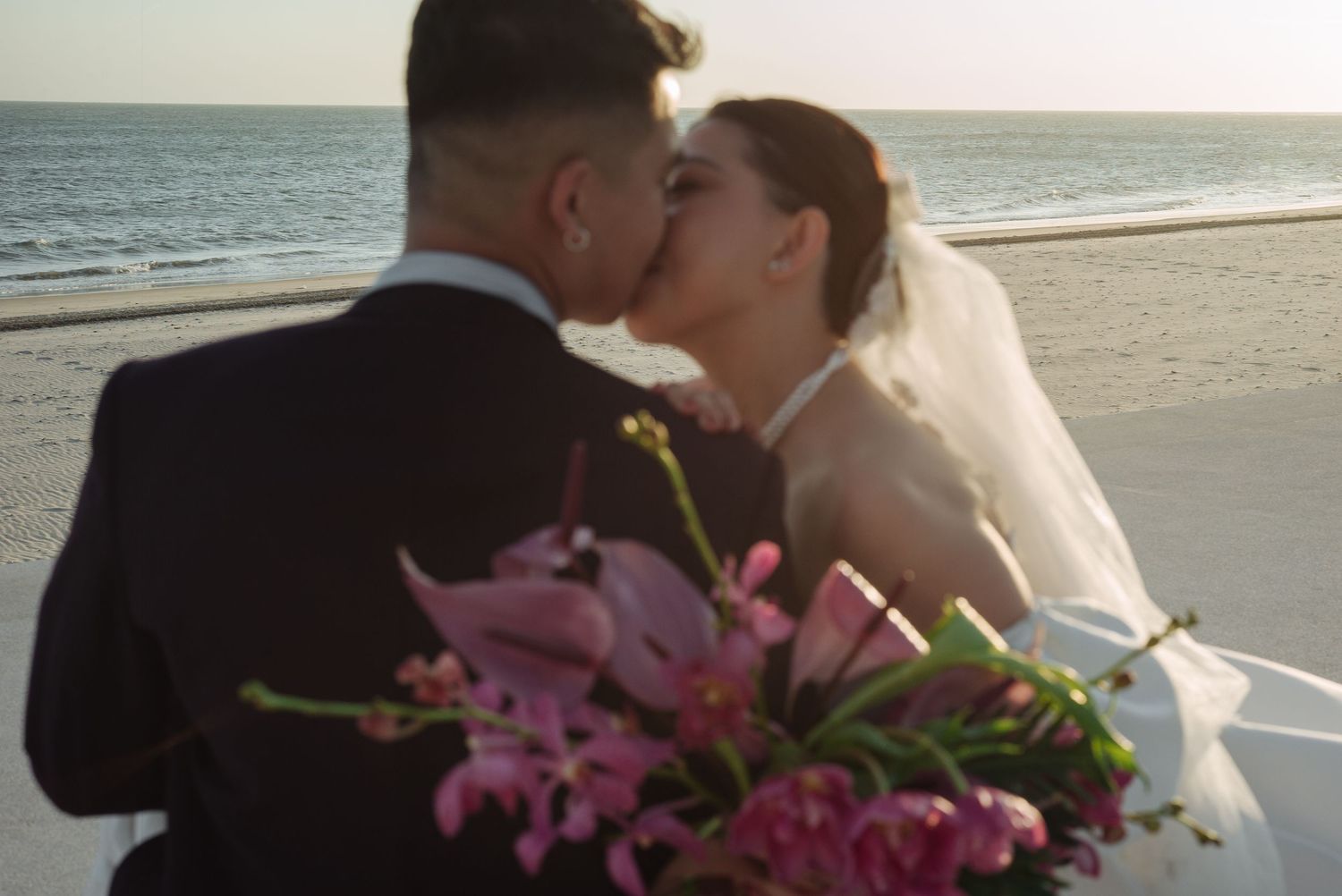 Bride and groom share romantic kiss on beach at sunset with pink tropical flower bouquet in foreground.