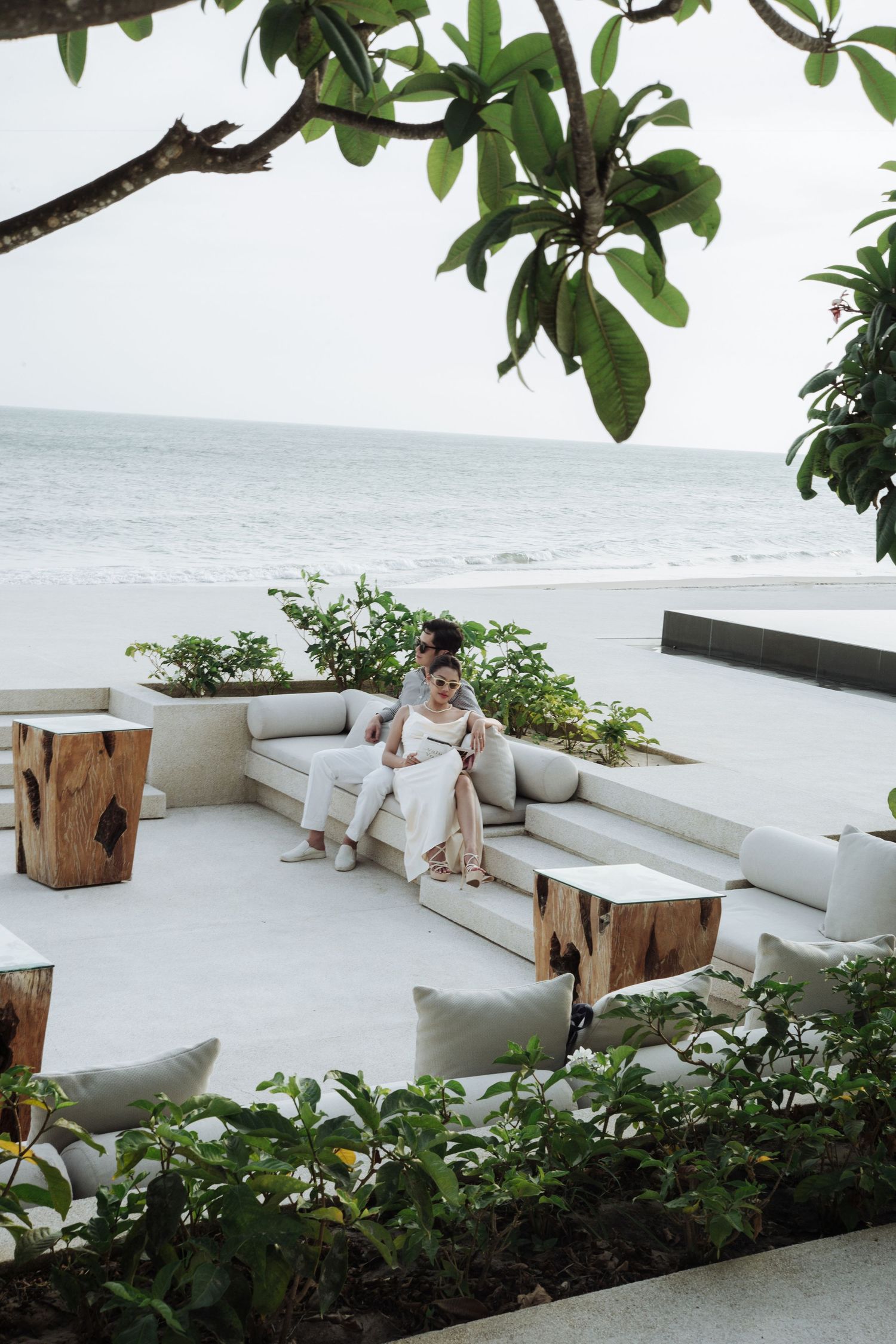 Couple relaxing on white outdoor sofa at beachfront luxury resort surrounded by tropical plants.