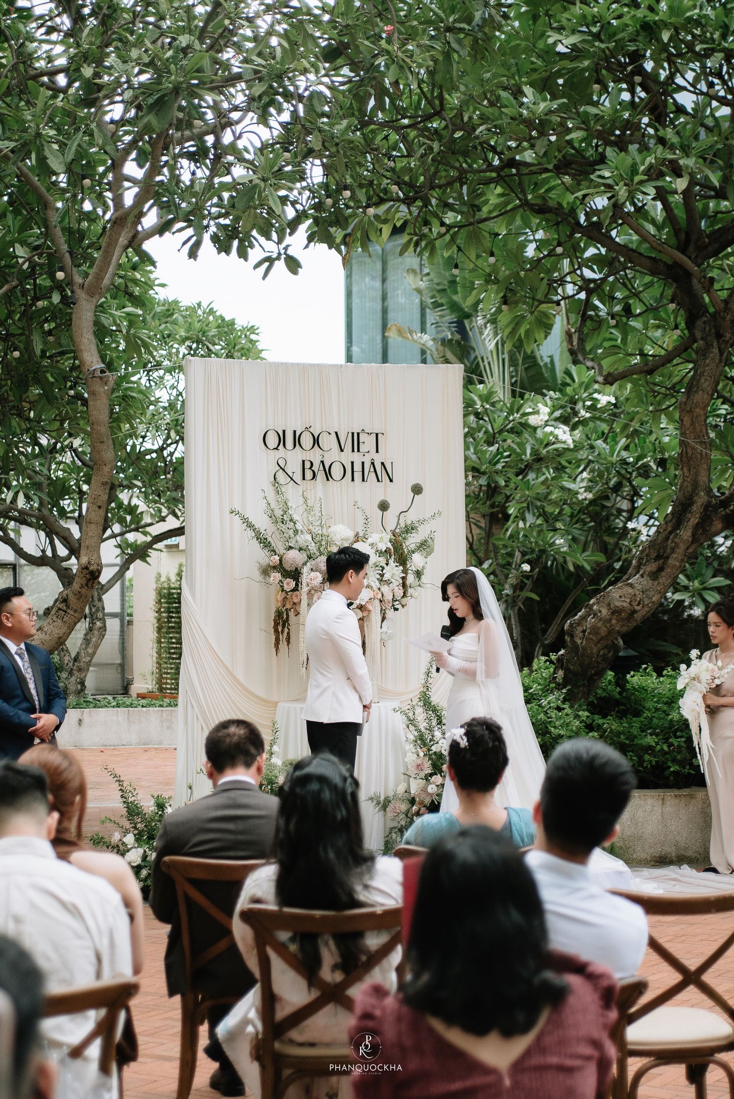 Wide shot of intimate outdoor wedding ceremony with guests seated before elegant white floral backdrop altar.