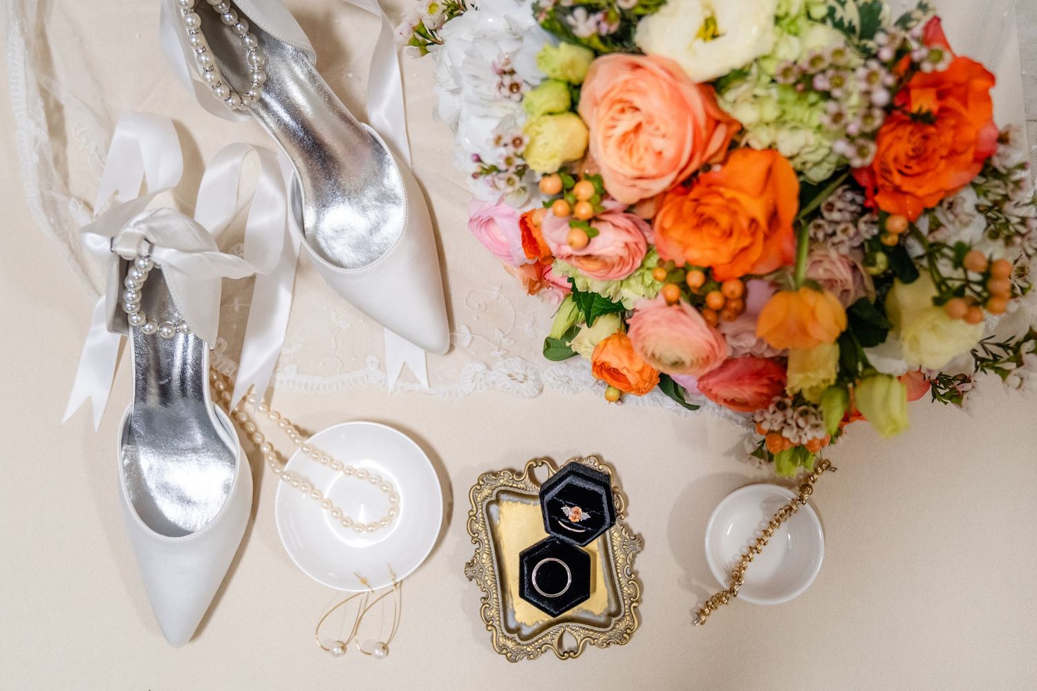 Wedding details flatlay with silver heels, vintage camera, and coral rose bouquet on white surface.