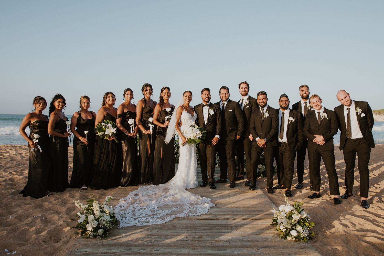 Wedding party in formal black attire poses on scenic beachfront.