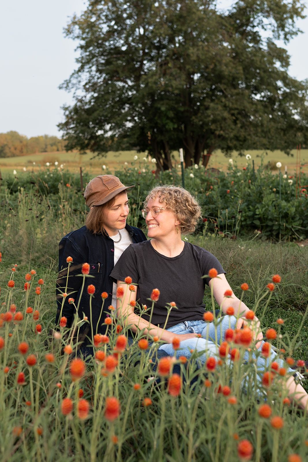 A couple sits together in a wildflower field with vibrant orange poppies blooming around them on a sunny day.