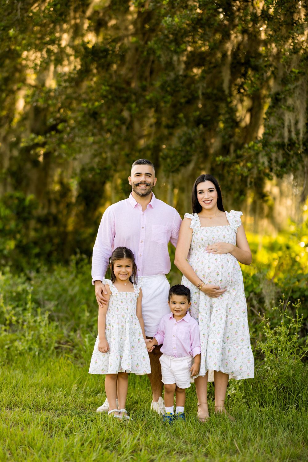 Family wearing coordinated white and pink outfits posing in a green meadow. Photo by Dani De La Rosa, DigitalMyst Photography, Wesley Chapel