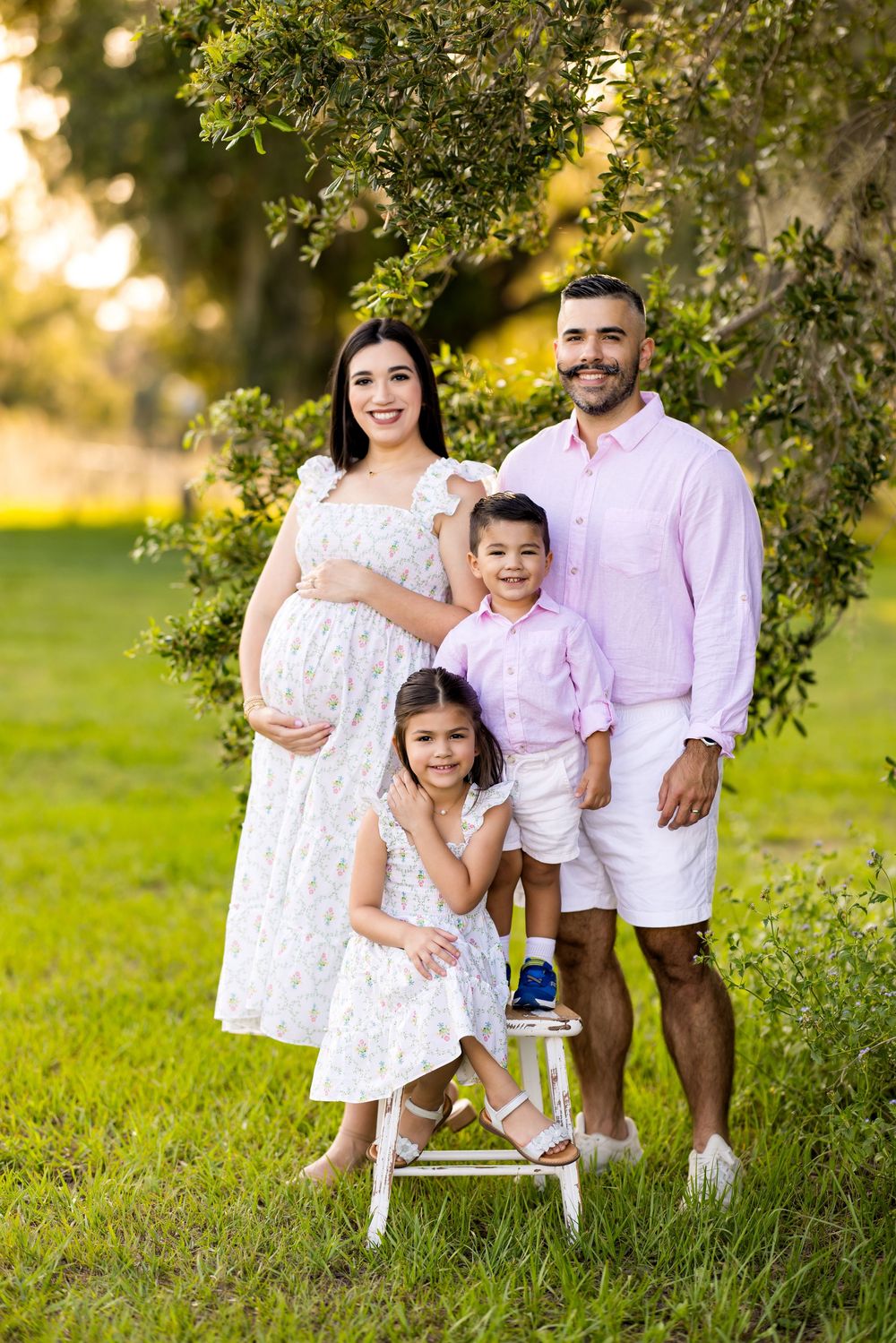 Family portrait in coordinated outfits sitting together in a sunlit garden setting. Photo by Dani De La Rosa, DigitalMyst Photography