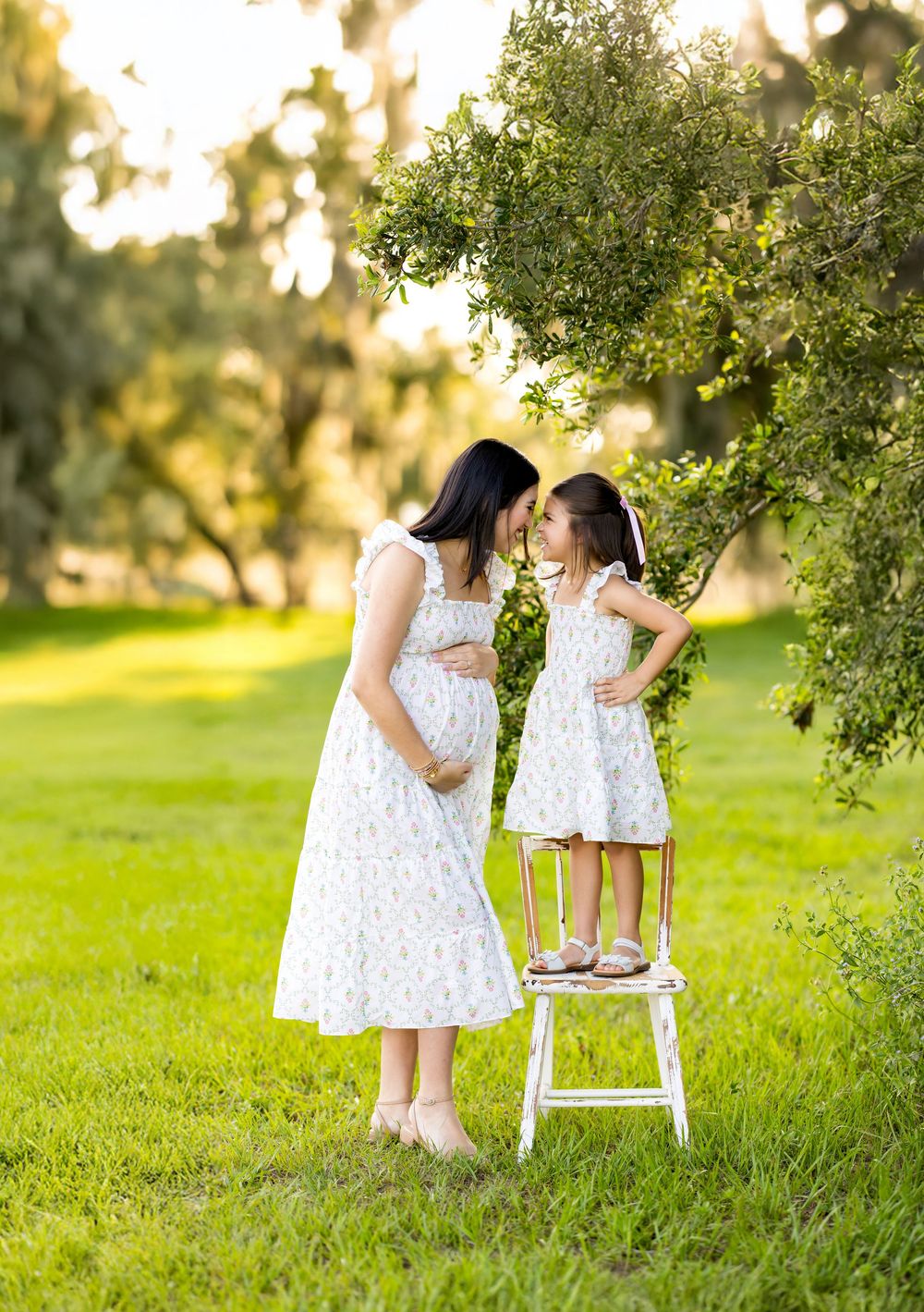 Two figures in matching white lace dresses share a tender moment in a sunlit grassy field. Photo by Dani De La Rosa, DigitalMyst Photography