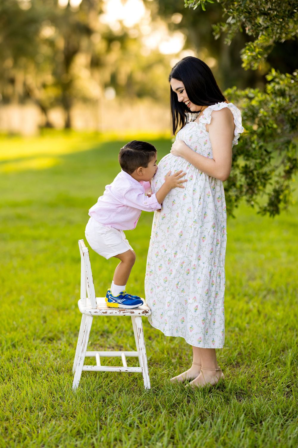 Young toddler stands on white chair reaching up toward figure in white lace dress in sunlit garden setting. Photo by DigitalMyst Photography