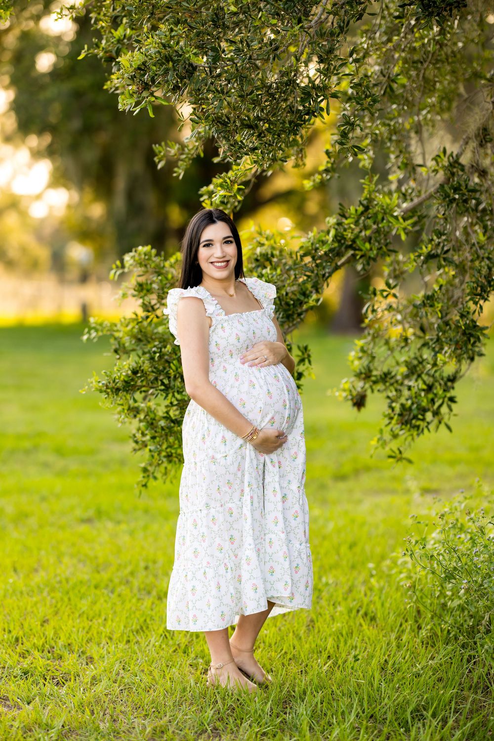 White lace maternity dress photographed against lush green garden backdrop at golden hour. Photo by Dani De La Rosa, DigitalMyst Photography