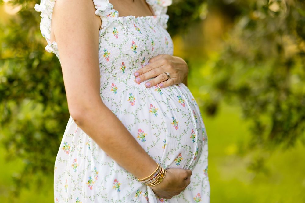 Close-up view of delicate white maternity dress with hands cradling baby bump in outdoor setting. Photo by Dani De La Rosa, DigitalMyst