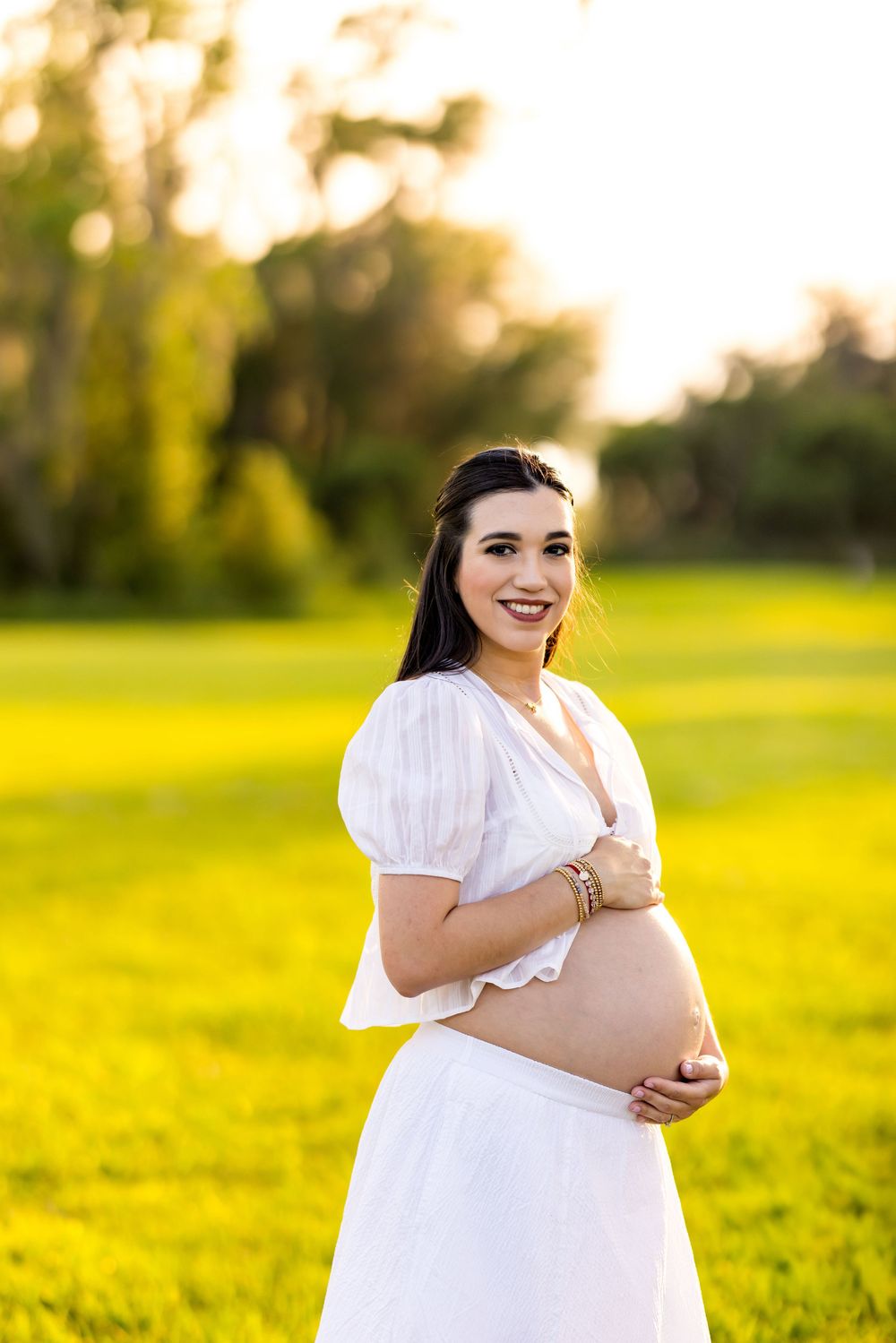 Maternity photo session with woman in white dress against golden sunset field backdrop. Photo by Dani De La Rosa, DigitalMyst Photography