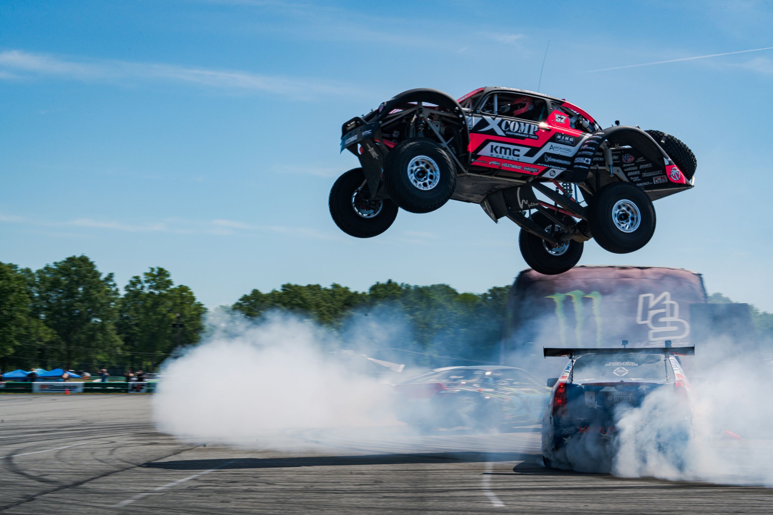 A red off-road truck soars mid-air over a smoking drift car on a racetrack under a clear blue sky.
