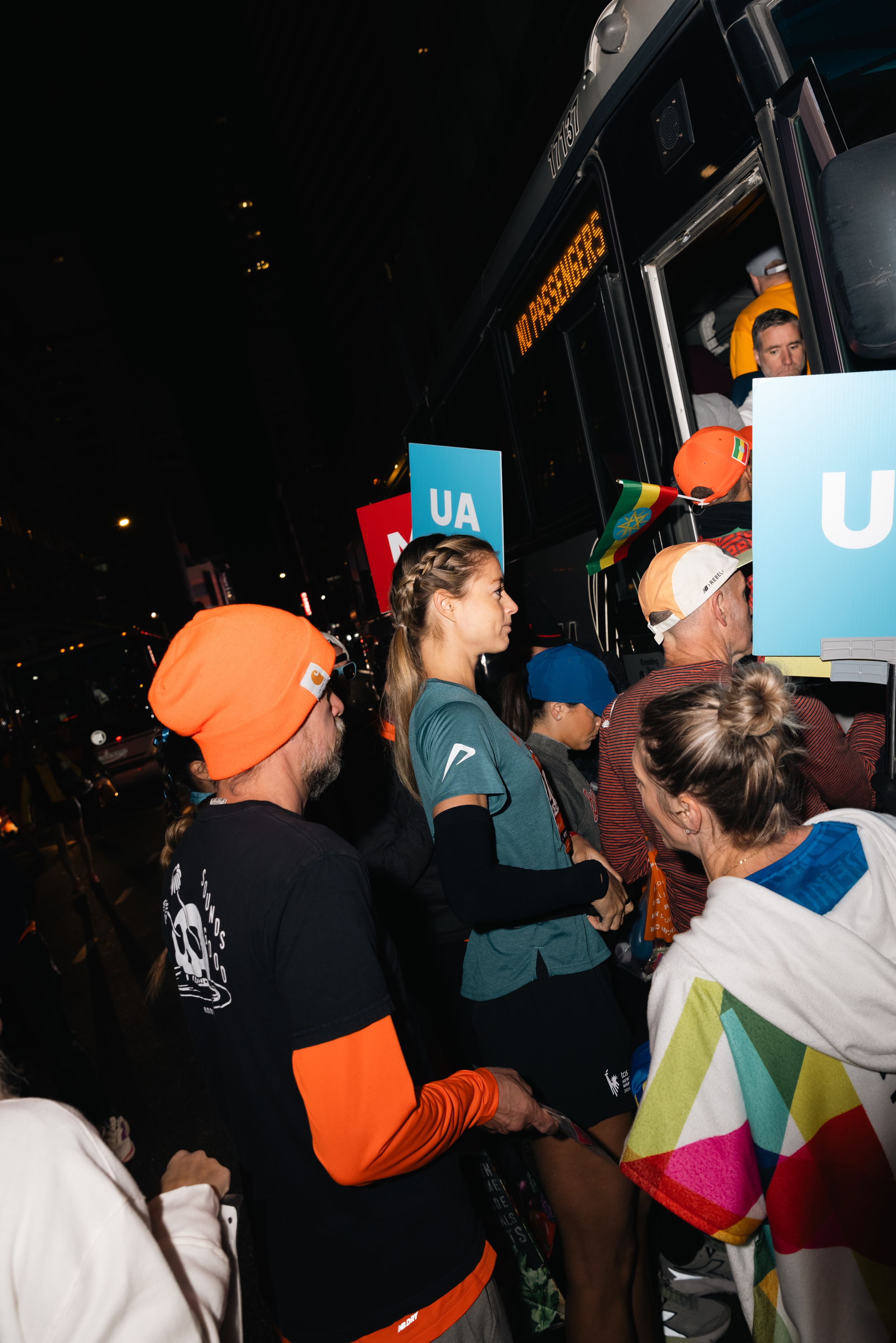 Crowd of marathon spectators and runners gathered around a bus at night.