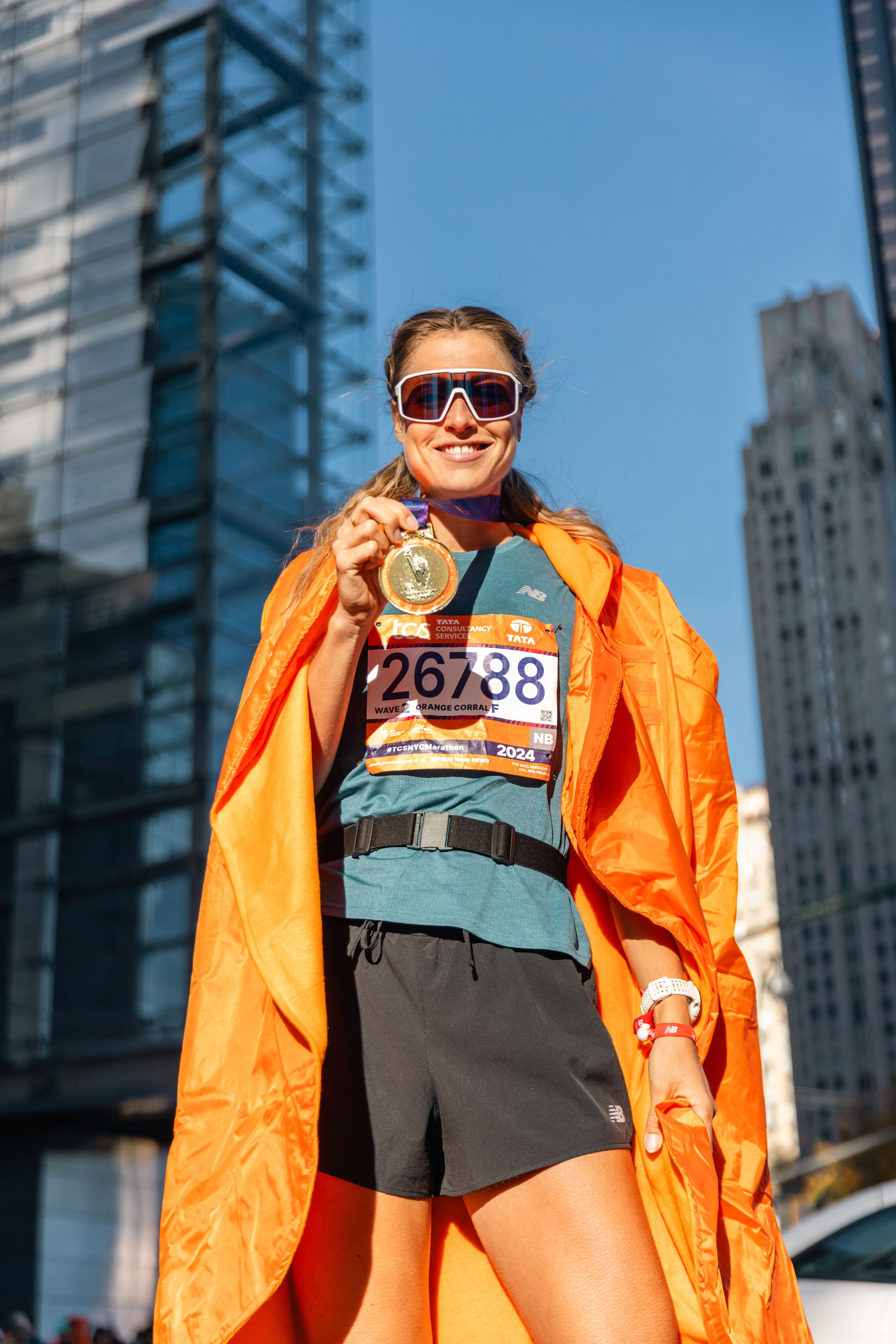 Runner in orange jacket displays marathon medal against urban skyline backdrop.