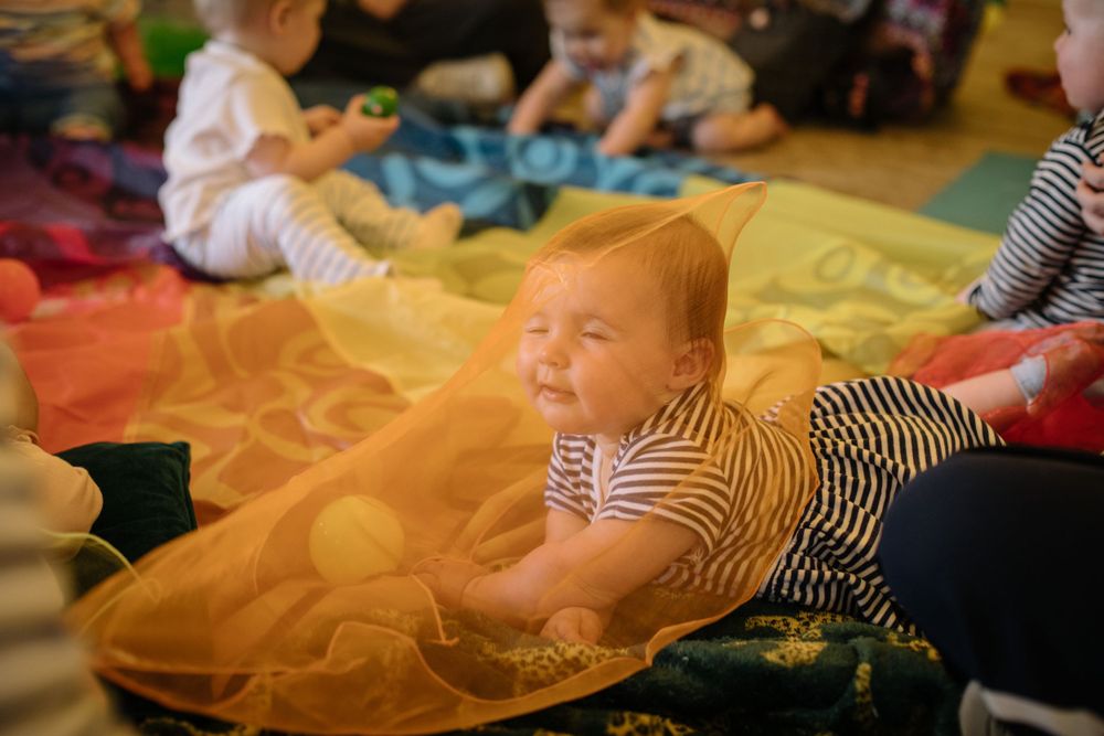 A baby in striped clothing lies on vibrant orange and yellow fabric during a group activity.