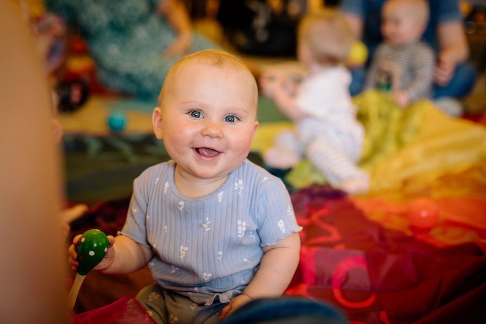 A baby in blue stripes sits on a colorful play mat during a sensory class activity.