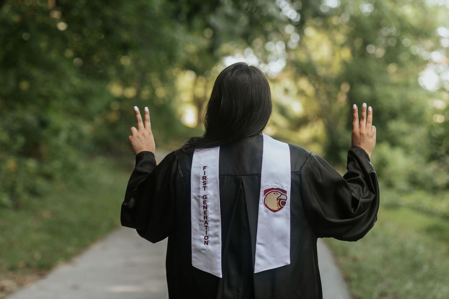 Graduate in black gown showing back with stole and raising hands on outdoor path.