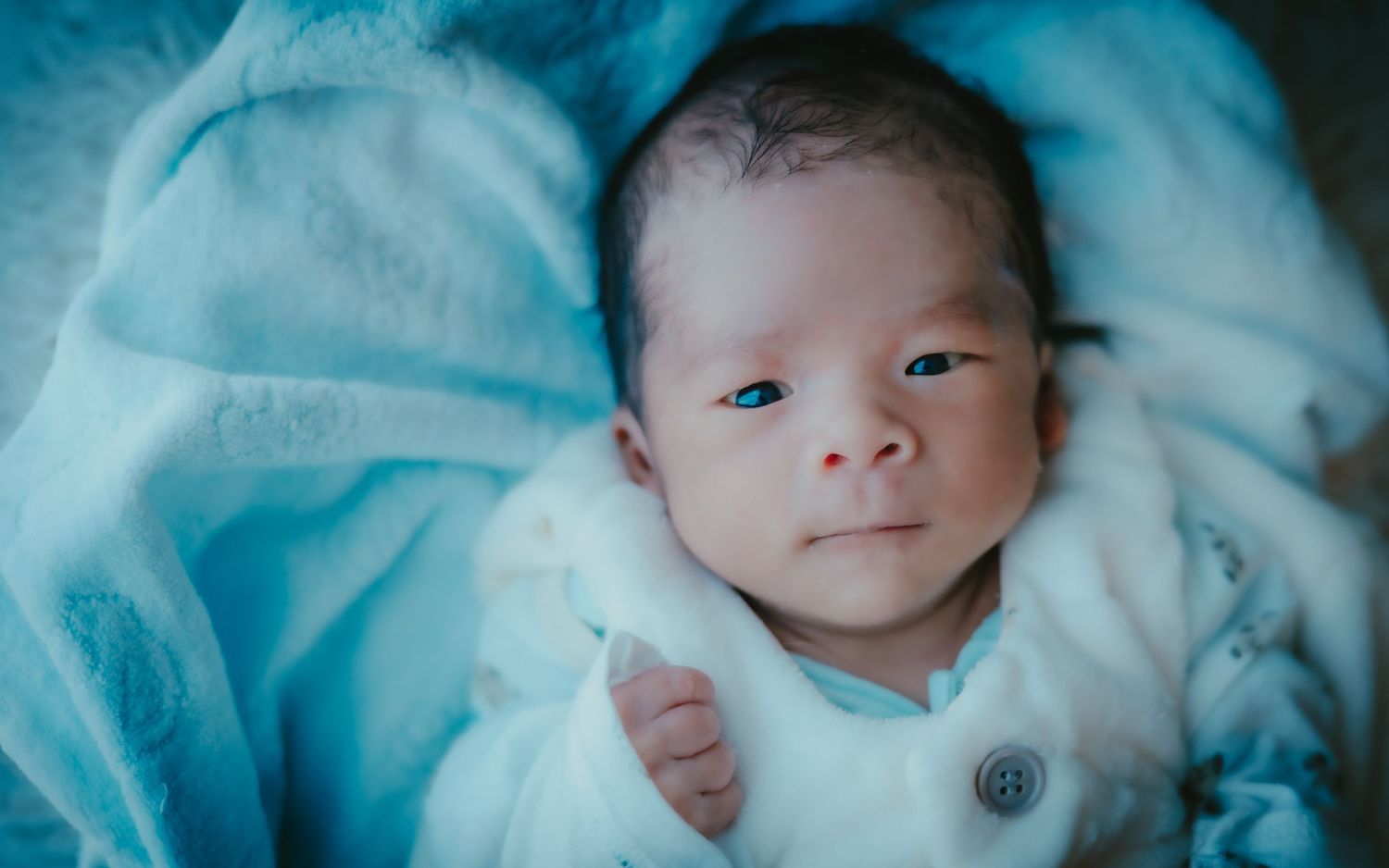 A newborn wearing a white knit outfit lies on soft blue blankets while gazing upward.