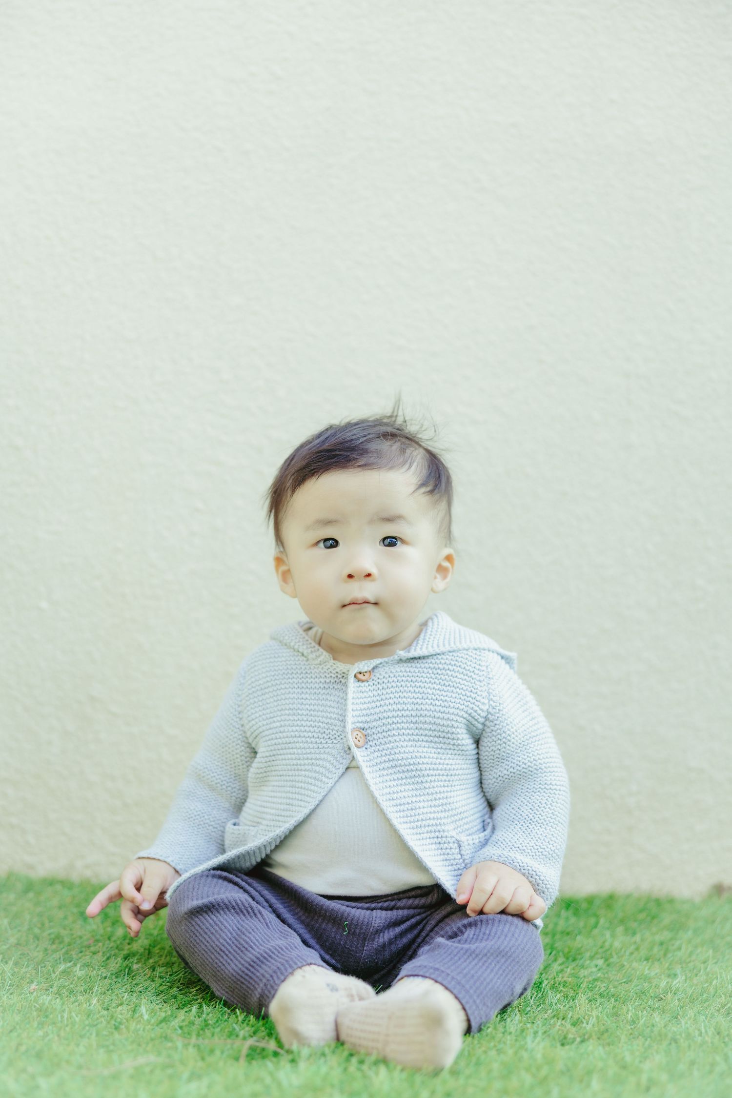 Baby in gray cardigan and navy pants sitting on grass against light wall.