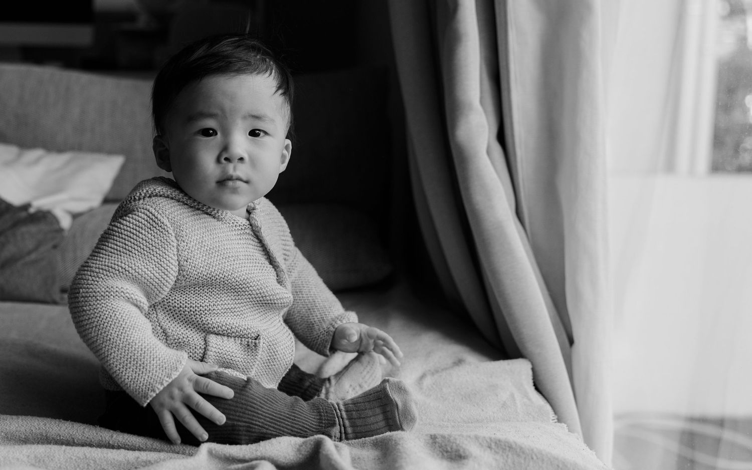 Black and white photo of toddler in light sweater sitting on bed.