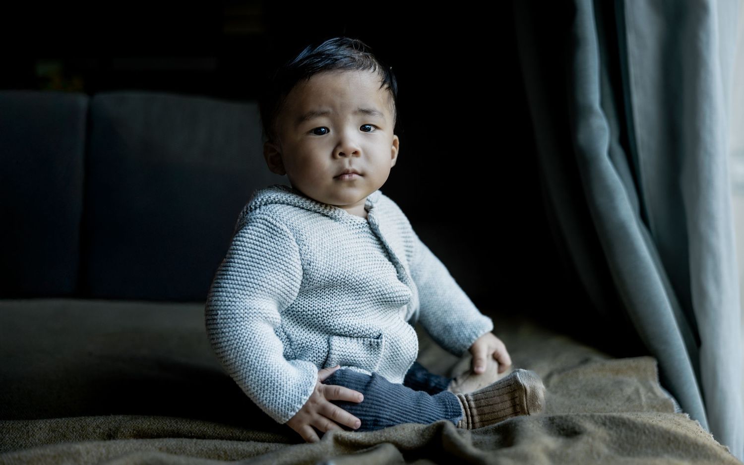 Toddler in gray sweater sitting with toy on dark couch.