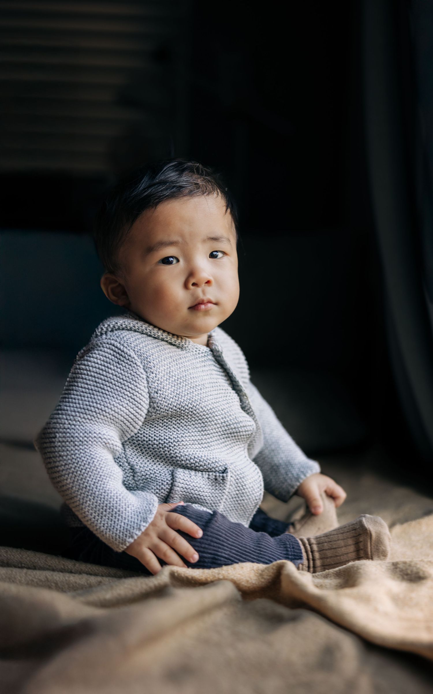 Baby in knit gray sweater sitting on textured blanket in moody lighting.