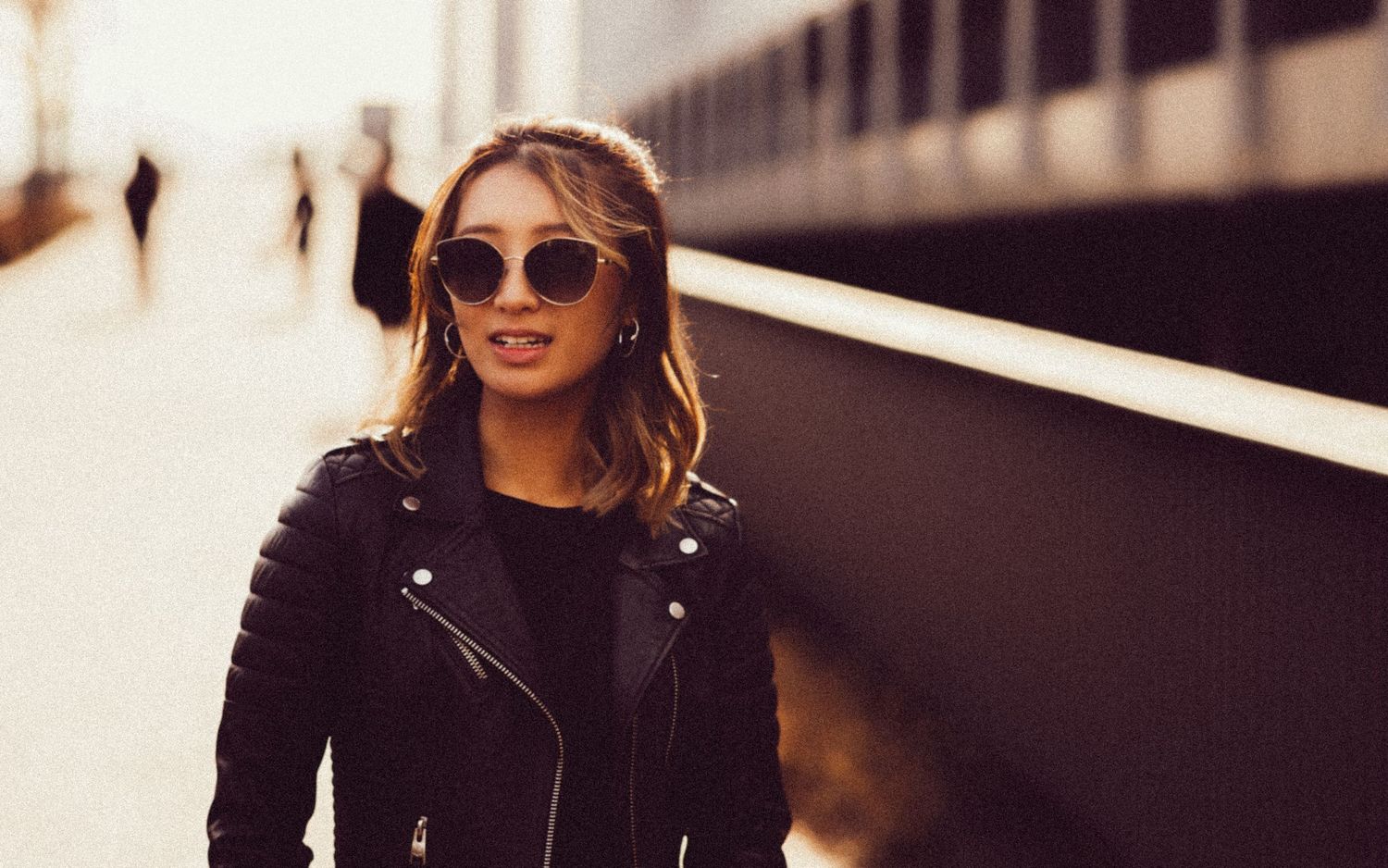 Person wearing black leather jacket and sunglasses poses outdoors in warm sunlight.
