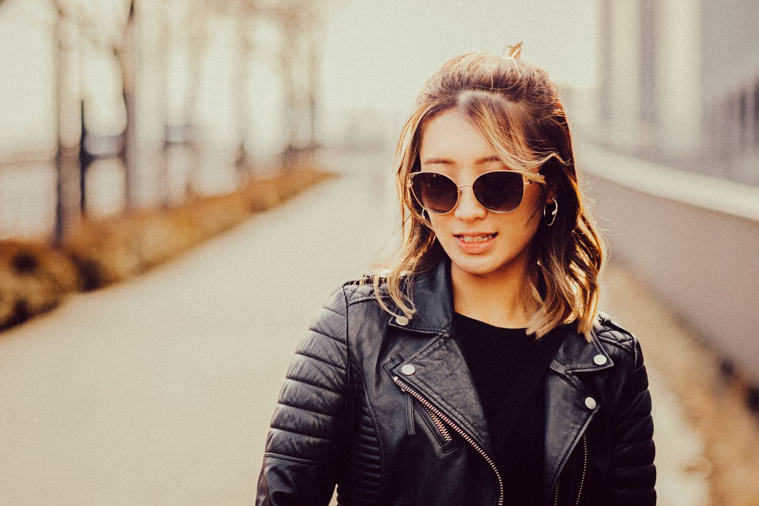 Person in black moto jacket and sunglasses smiles while posing on urban walkway.