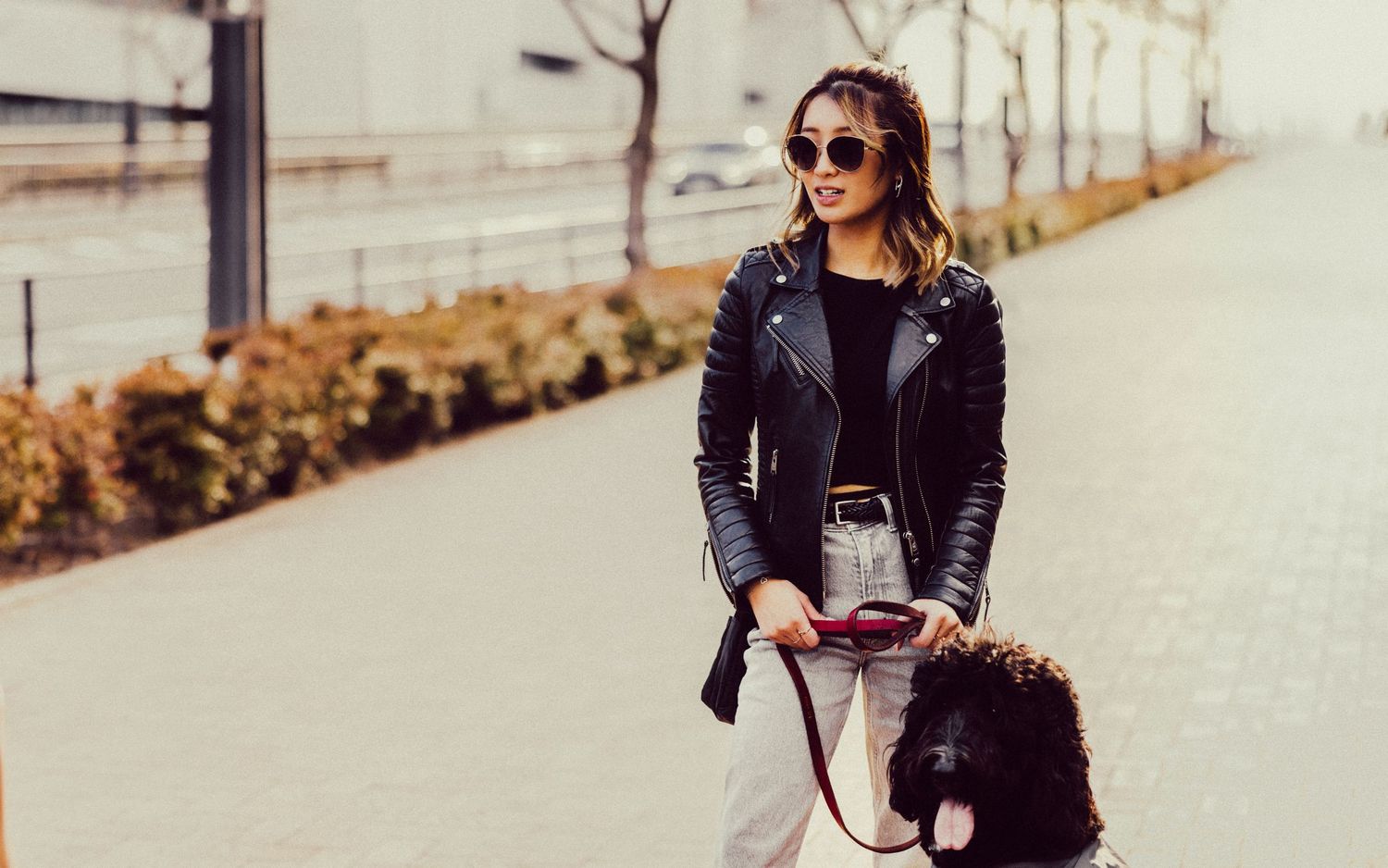 Person in black leather jacket and sunglasses poses with handbag on urban pathway.