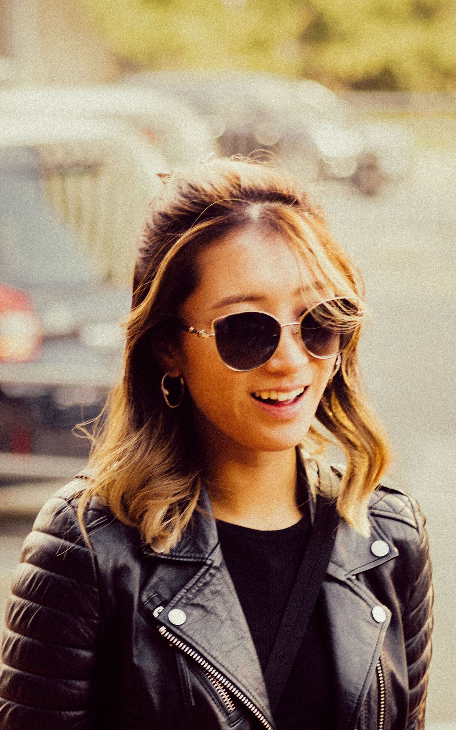 Person wearing black leather jacket and aviator sunglasses smiles warmly outdoors.
