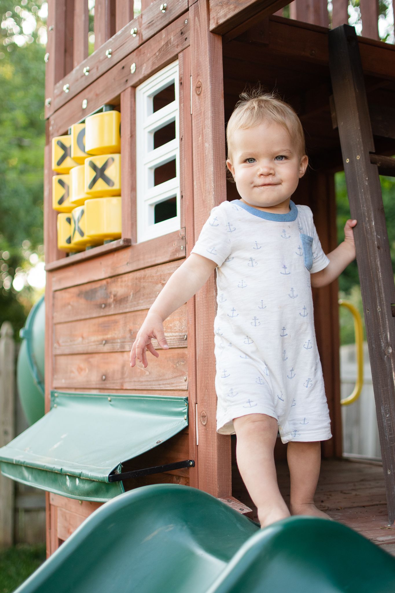A toddler stands on a wooden playhouse structure with a green slide, wearing a light colored romper.