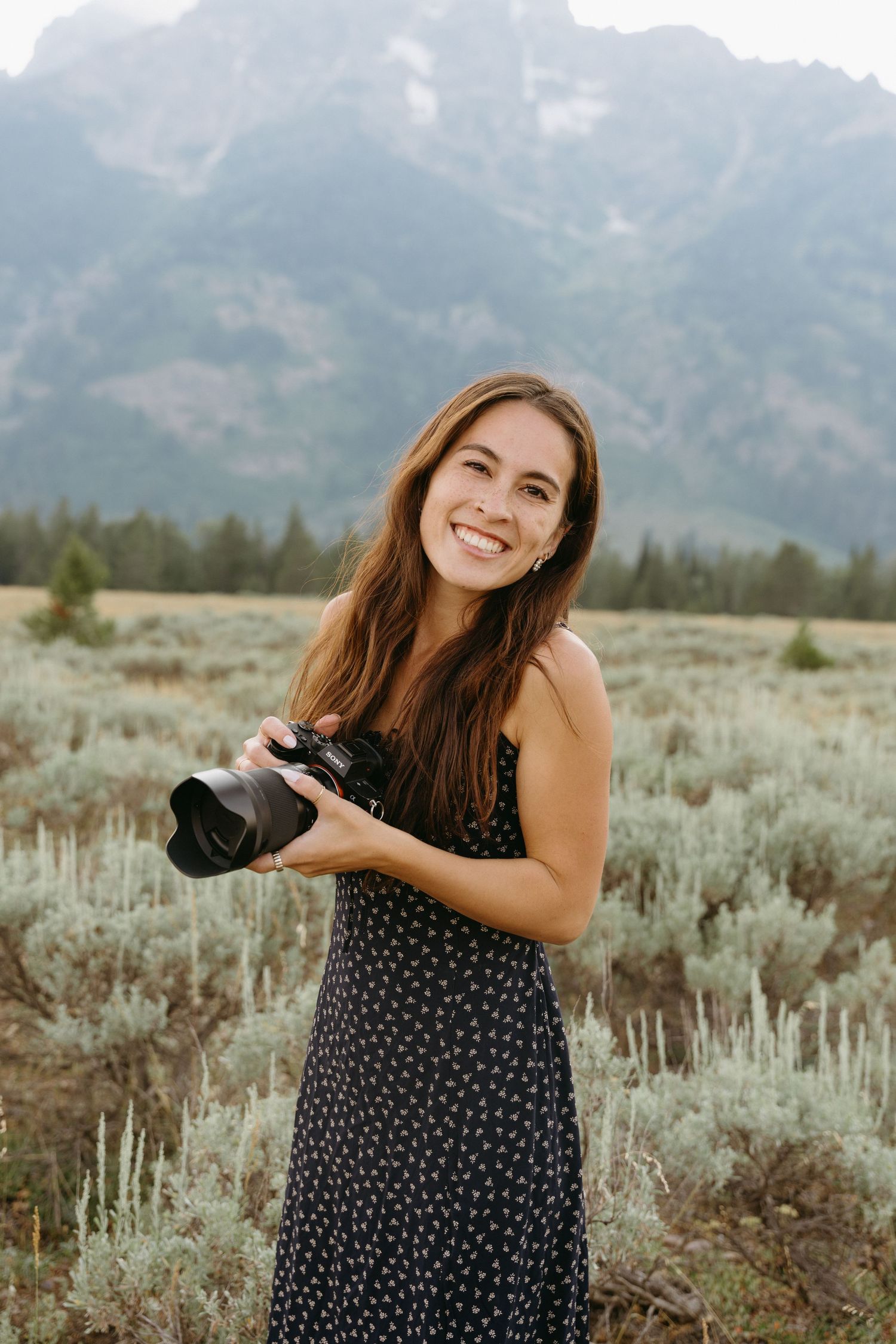 A photographer in a polka dot dress holds a camera while standing in front of mountain peaks and sagebrush.