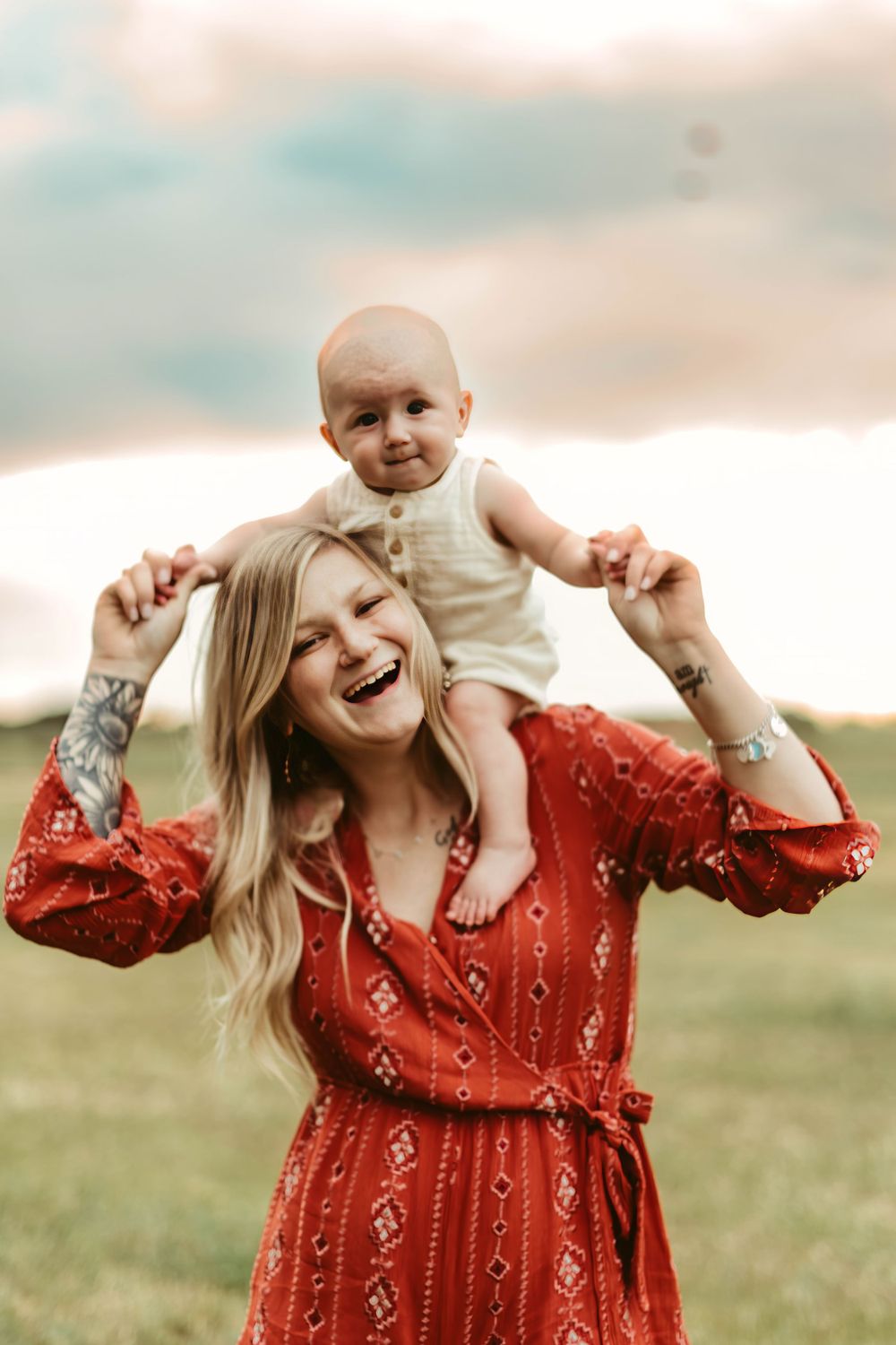 A baby sits on shoulders while someone in a red dress laughs in an outdoor field setting.