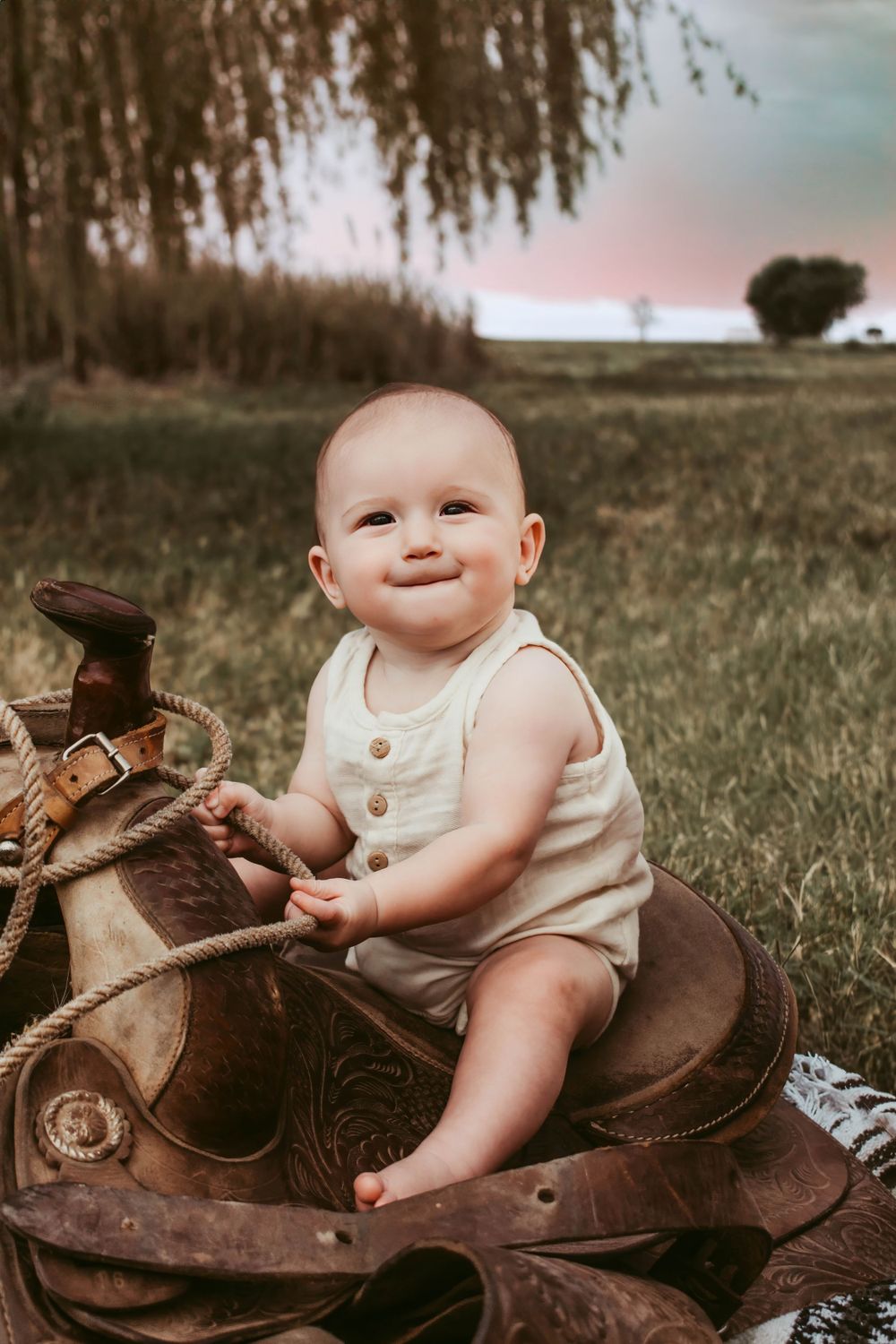 A smiling baby sits on a vintage leather saddle in an outdoor setting.