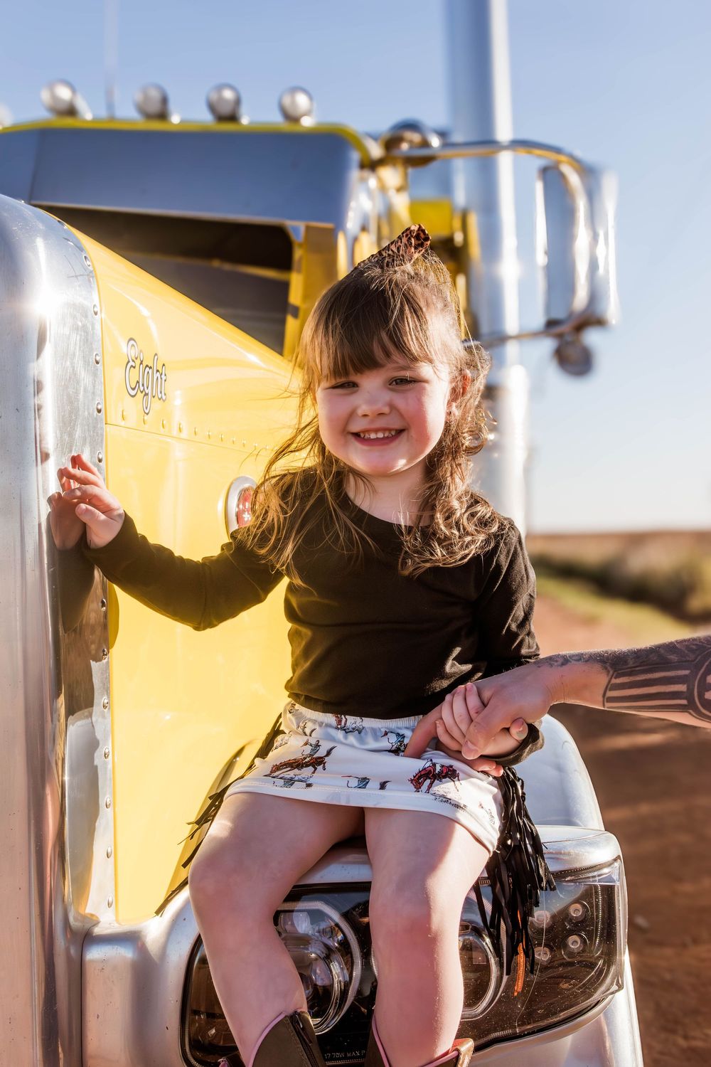 Child smiles while sitting on semi truck steps during golden hour photoshoot.
