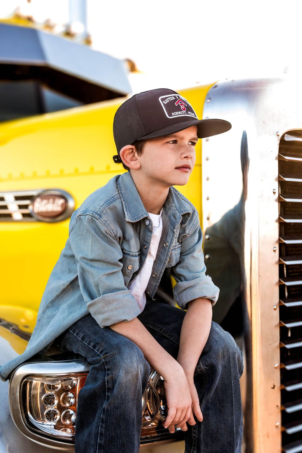 Child model sits on yellow vehicle wearing denim jacket and baseball cap during daytime shoot.