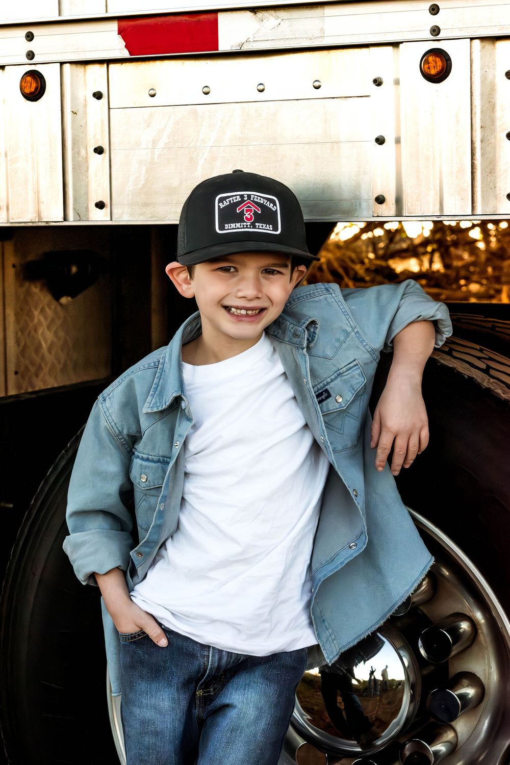 Young person in denim jacket and baseball cap poses casually against vehicle in outdoor photoshoot.