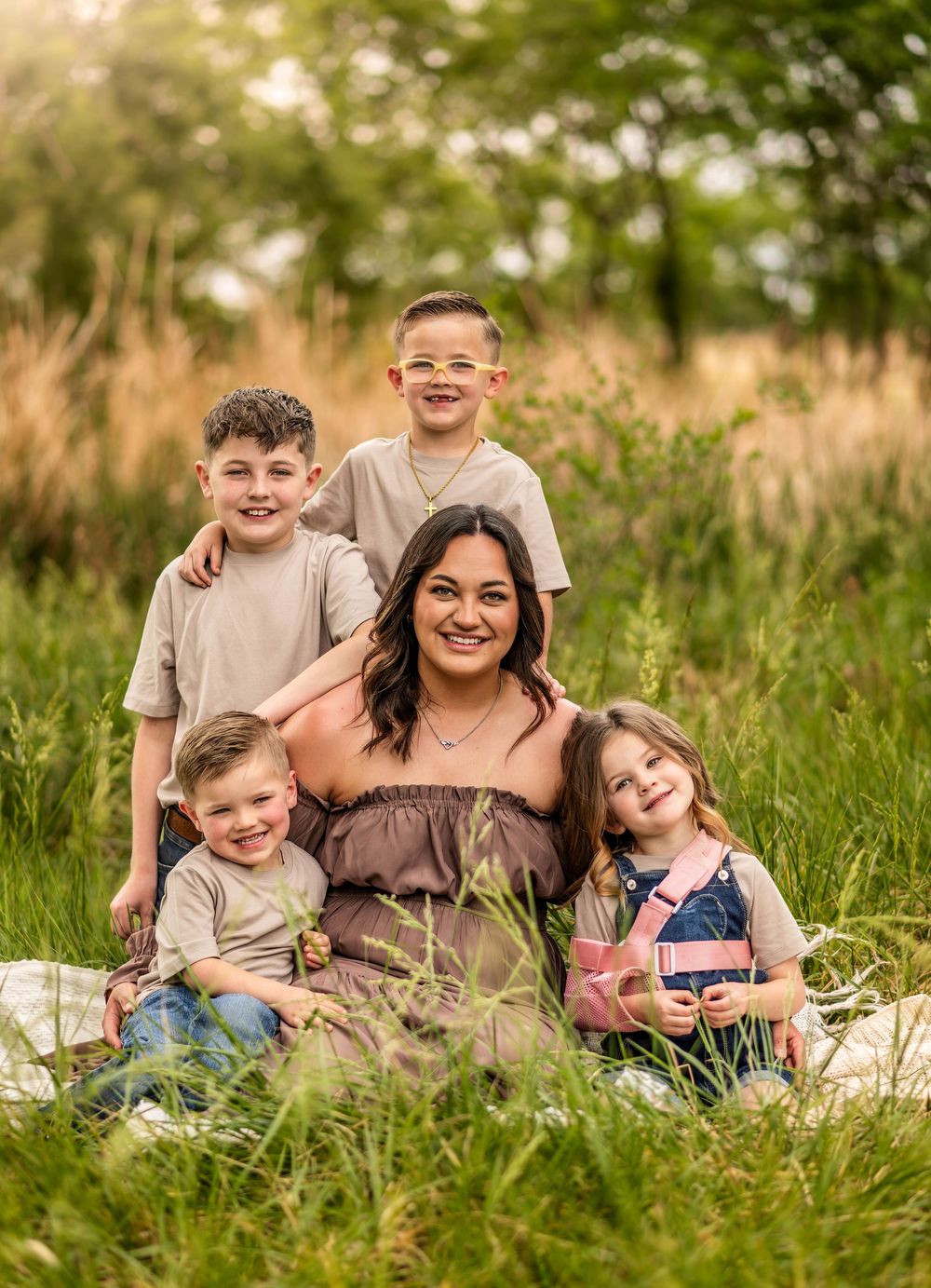 A smiling woman sits in a lush green field with four young children around her for a family portrait.