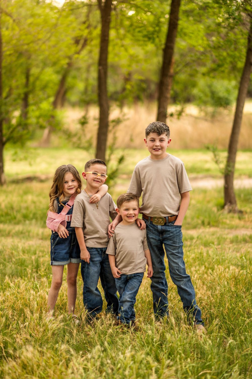 Four children in matching tan shirts and jeans pose together smiling in a lush green outdoor park setting.
