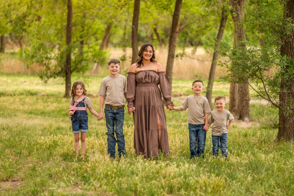A mother in a brown dress holds hands with four young children in a lush green park setting.