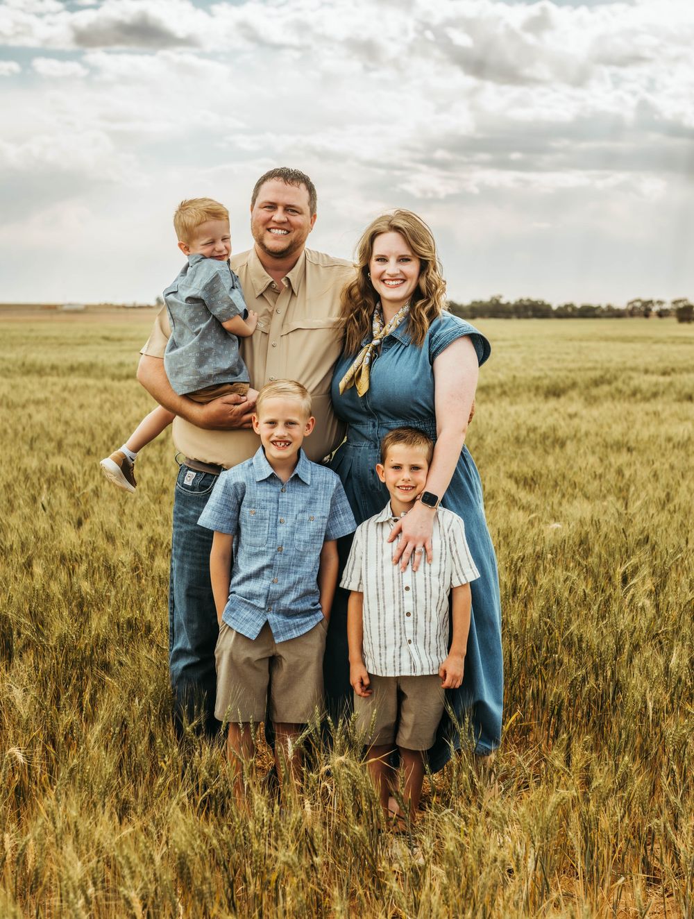 Happy family of five posing in a golden wheat field, parents with three young boys smiling outdoors.